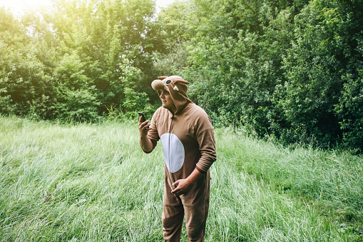 Young man is standing in the forest in cosplay costume of a cow. Guy in the funny animal pyjamas sleepwear is holding smartphone in the nature. Halloween ideas for party.