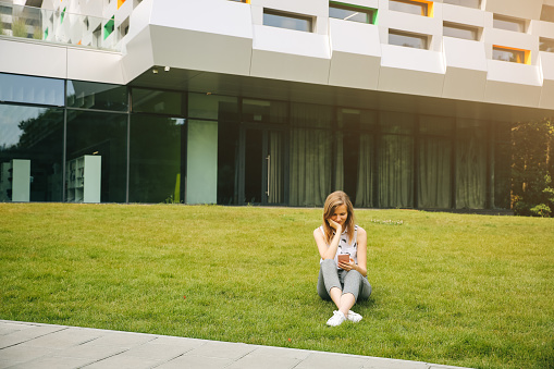 Beautiful hipster girl is sitting near modern architecture building. Stylish business woman looking in the smartphone on green grass lawn. Student surfing in networks. Lifestyle relax. Office worker.