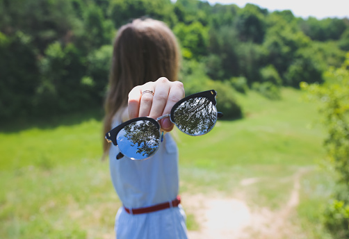 Young hipster girl is travelling in the mountains. Woman in modern dress is holding fashion sunglasses in hands. Trees and sky reflection. Freedom style concept. Nature in summer. Holiday vacations.