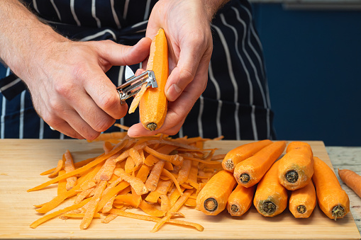 male chef hands peeling a bunch of carrots with a food peeler in front of a striped cooking apron in the kitchen. preparing fresh vegetables on a wooden cutting board for cooking a healthy meal.