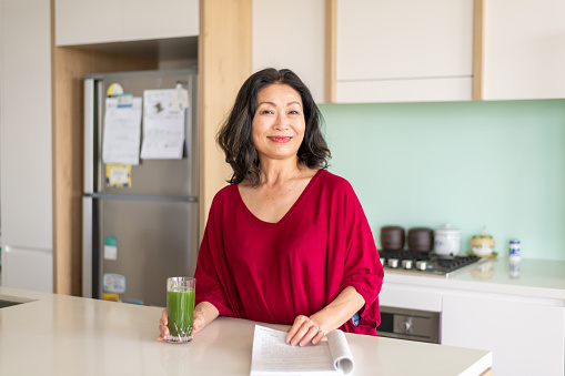 Attractive woman of asian ethnicity reading paper and drinking vegetable juice. Wearing a red sweater, looking directly at the camera and smiling.