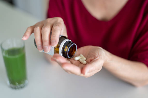 Keeping on top of good health Close up shots of hands pouring out some vitamins. Green smoothie in a glass in the background. Woman is wearing a red coloured sweater. nutritional supplement stock pictures, royalty-free photos & images