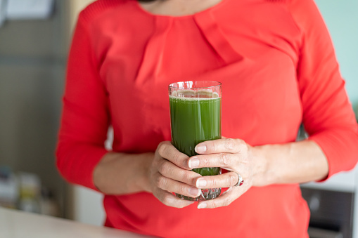 Close up shot of two hands holding a glass of green smoothie. The woman is wearing a bright red top.