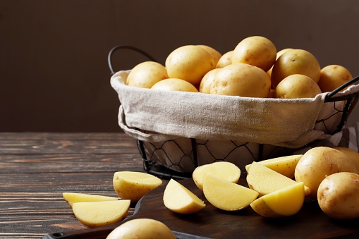 Basket full of fresh, young potatoes board, towel and knife on wooden background, top view