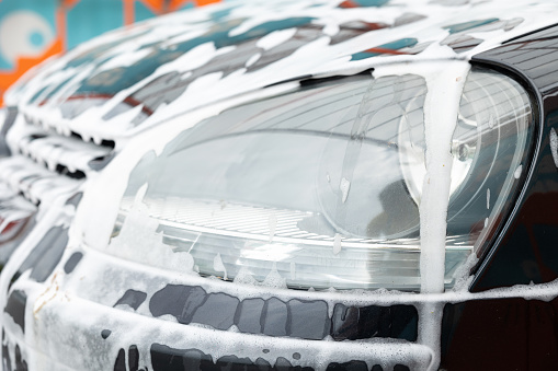 The headlight and soot of the car are coated with white detergent. Close-up. The car covered in white foam at a self-service car wash.
