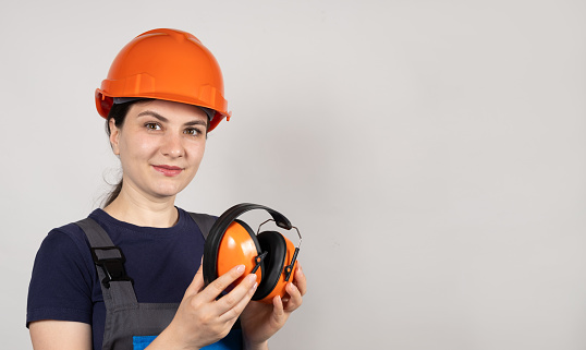 Female construction worker in protective helmet holding earmuffs on white background