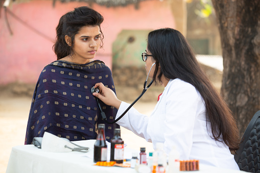 Indian female doctor checking young adult girl patient heartbeat with stethoscope at village outdoor, woman getting examine by medical person, Rural India healthcare concept