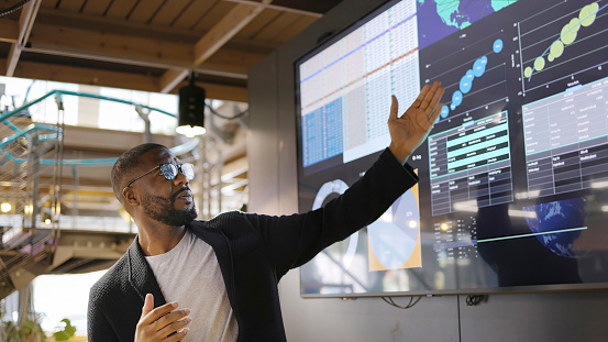 Stock image of a Black man conducting a seminar / lecture with the aid of a large screen. The screen is displaying graphs & data associated with movie clips of the earth.