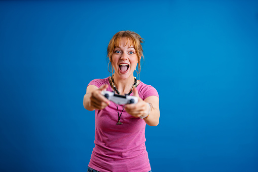 Portrait of a beautiful young woman standing in front of a bright blue background and playing a video game on a joystick.