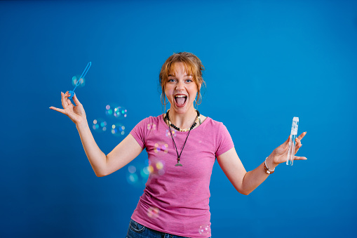 Portrait of a beautiful young woman blowing bubbles in front of a bright blue background.