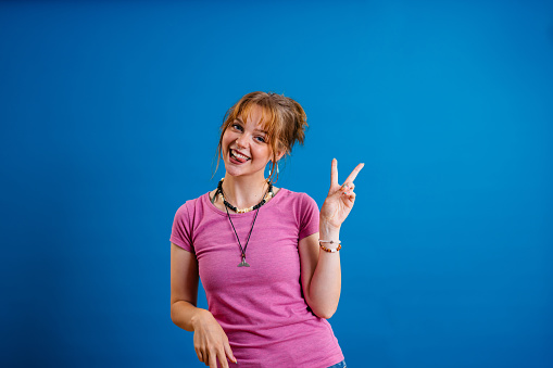 Portrait of a beautiful young woman holding up a peace sign while standing in front of a bright blue background.