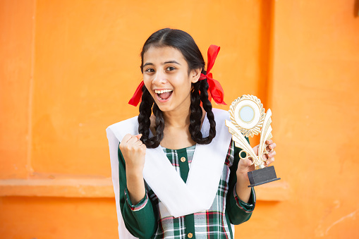 Young beautiful happy indian school girl celebrating victory with trophy,Cheerful excited braided female holding winning prize against orange background, skill india.