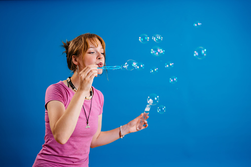 Portrait of a beautiful young woman blowing bubbles in front of a bright blue background.