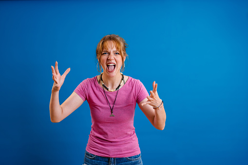Portrait of an angry young woman standing in front of a blue background and shouting.