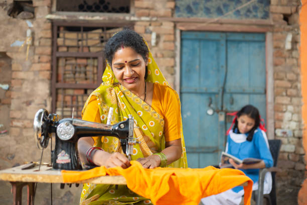 donna indiana rurale che usa la macchina da cucire mentre la sua giovane figlia studia dietro di lei. - sewing women tailor teenage girls foto e immagini stock