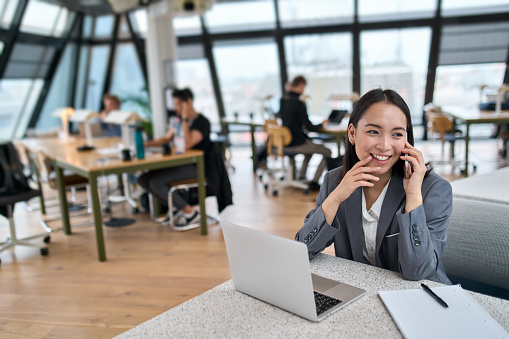 Young happy Asian business woman wearing suit working in big modern office. Smiling female korean company manager, professional worker entrepreneur or employee using laptop talking on the phone.