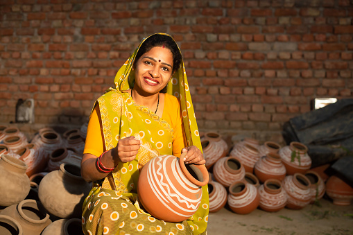 Portrait of happy traditional Indian woman potter artist painting and decorating design on clay pot for sale, handicraft, skill india.