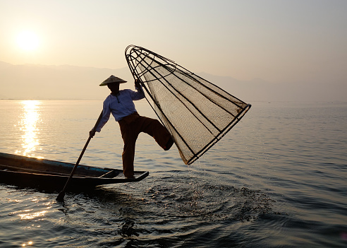 Inle, Myanmar - Feb 15, 2016. A Burmese man using the unique methods of rowing and catching fish on Inle Lake, Myanmar. Inle is one of the highest lake at an elevation of 2,900 feet (880m).