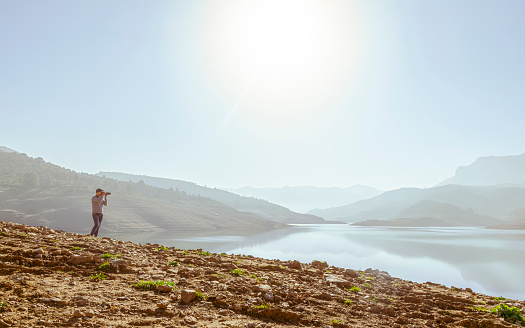 A man taking photograph in Bouzegza Keddara,  Boudouaou, Algeria. Brown soil, bush and shiny sunlight in a blue sky reflected in water.