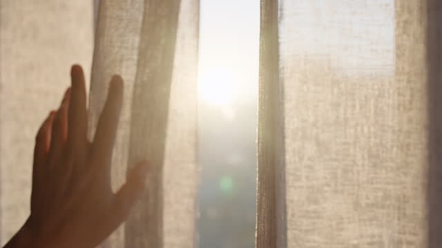 Window, sunrise and curtain with hand of person in bedroom for wake up, morning and light. Silhouette, relax and texture with closeup of woman feeling fabric at home for sunshine, peace and calm