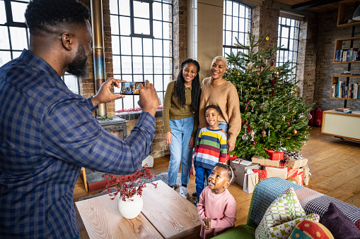 Bearded Black man holding smart phone and capturing holiday memory of woman standing with children aged 2 to 12, all casually dressed and smiling at his camera.