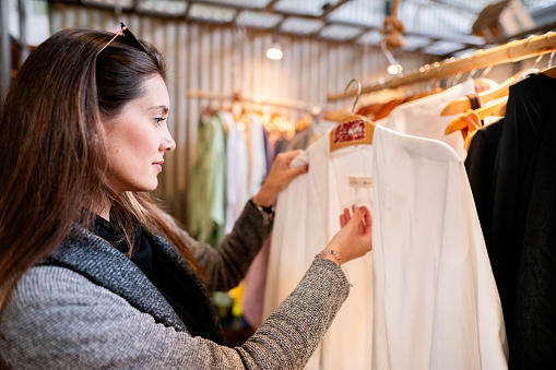 Stylish young woman looking at a tag while browsing clothing hanging on a rack in a market stall