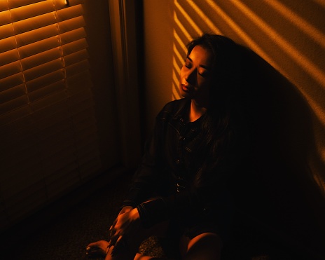 A young Southeast Asian female is exhaustedly leaning against a wall in a dimly-lit room