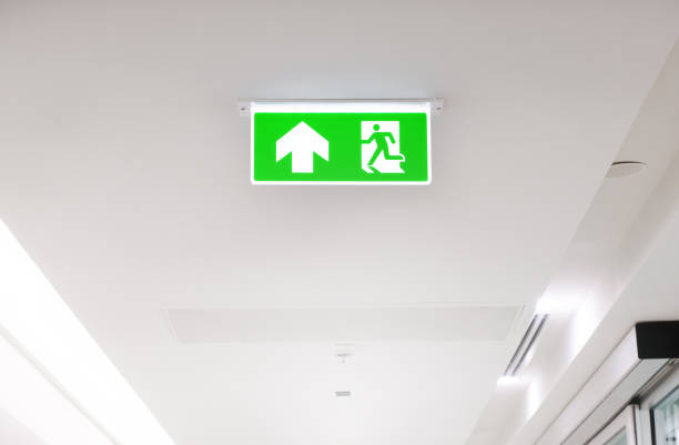 istock Lighted glowing green emergency exit signs in an office hallway with arrows pointing the way out of the building. 1614436385