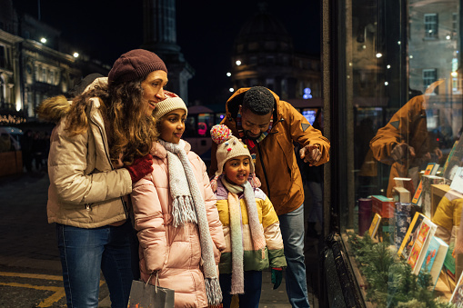 Side view of two parents standing outside of a store window with their two young daughters, they are shopping for Christmas presents and they're wrapped up in warm clothing on a cold December night.