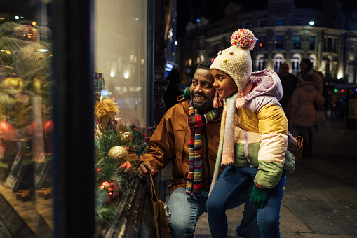 Side view of a father standing outside of a store window with his young daughter, they are shopping for Christmas presents and they're wrapped up in warm clothing on a cold December night.