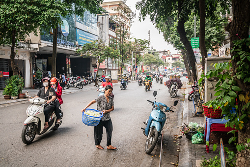 Hanoi, Vietnam, November 13, 2022: An elderly woman carrying a basket of vegetables navigates scooter traffic while crossing a busy side street in Hanoi, Vietnam