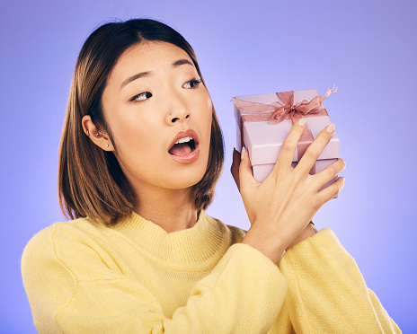 Surprise, gift and box with face of asian woman in studio for birthday, celebration and curious. Thinking, wow and party with female person and present on purple background for giveaway and package
