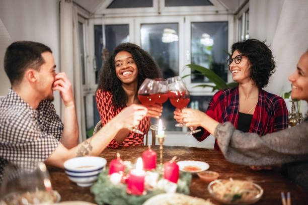 man and three woman around wooden table clinking glasses at christmas party four people sitting around wooden dining table with food and advent wreath and clinking wine glasses advent candle wreath christmas stock pictures, royalty-free photos & images