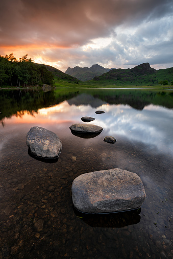 Dramatic sunset reflections in lake with mountains. Rocks in foreground. Rugged British landscape in rural open countryside. Blea Tarn, Lake District, UK.