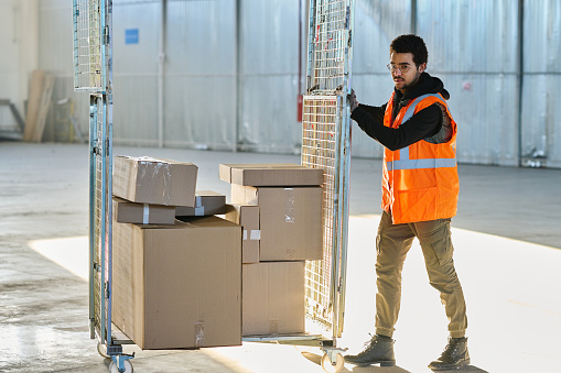 Young worker of warehouse in workwear pushing cart with stack of packed cardboard boxes containing spare parts for industrial equipment