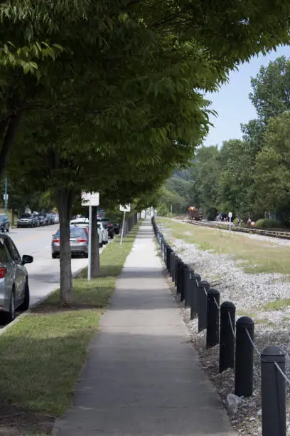 A sidewalk with trees, train tracks, guardrails, and cars right off the Ohio river in Cincinnati