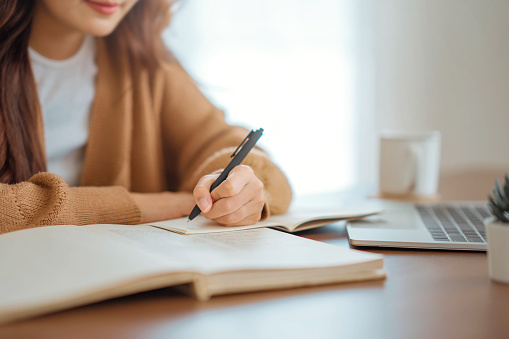 Close up - Hand of woman writing in spiral notepad placed on wooden desktop with various items at home. Copy space