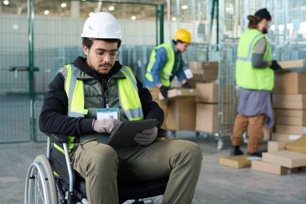 Young serious worker of warehouse sitting in wheelchair and using tablet stock photo