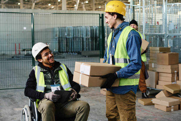 Happy young engineer in wheelchair looking at colleague carrying boxes stock photo