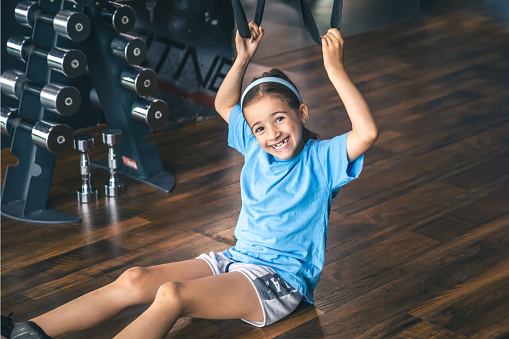 Little girl exercising indoors using trx fitness straps for working with his own weight, doing squats or training upper body.