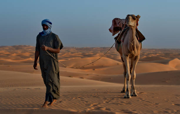 A Tuareg man dressed in traditional blue clothing sits on a sand dune in the Sahara desert. A portrait in which the sharpness is on the hands and the face is blurry.
