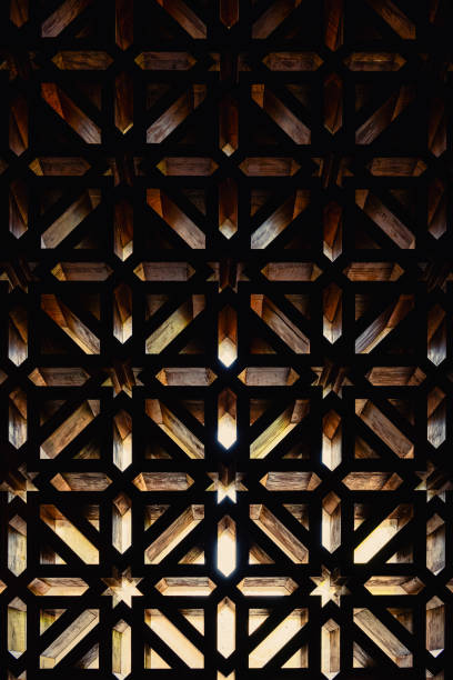 Vertical photograph taken from the interior of the Mosque-Cathedral of Cordoba, Spain. The shot captures light streaming through the gridded window, creating a dramatic pattern in warm tones. Taken with a slight upward perspective, the image emphasizes the window's geometric designs in a symmetrical composition, making it an ideal wallpaper or texture and a representative image of Cordoba.