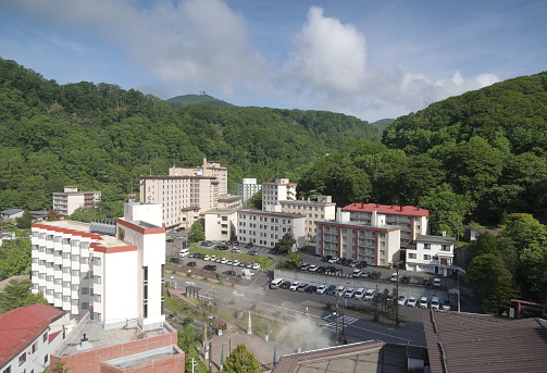 Noboribetsuonsencho, Japan - June 8, 2023: Hotels line Route 350 in the hot springs town of Noboribetsu Onsen. Foreground shows the rising steam in the Sengen Park plaza lined with colorful maces of the oni, or demons of the nearby Jigokudani, Hell Valley. Spring morning in the Central Area of Hokkaido Prefecture.