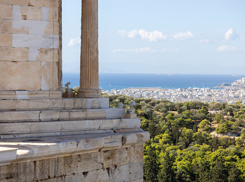 Temple of Athena Nike at Propylaia, monumental ceremonial gateway to the Acropolis of Athens, Greece. In the distance, aerial view of the city and sea with port of Piraeus