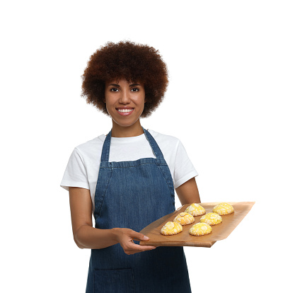 Happy young woman in apron holding board with cookies on white background