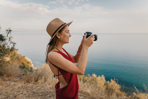 A young woman wearing a hat enjoying her vacation at the seaside and taking photographs with a camera on a lovely summer day