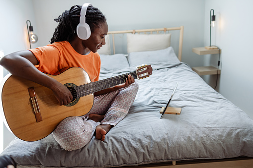 Young African American woman in bed, watching online tutorials while playing a guitar