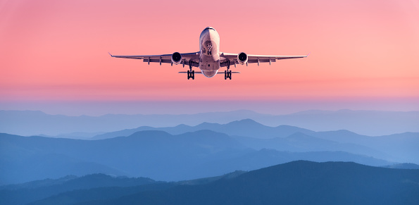 Plane is flying in pink sky at sunset. Landscape with passenger airplane over mountains ranges in fog, red sky at twilight. Aircraft is landing. Business. Aerial view. Transport. Private Jet
