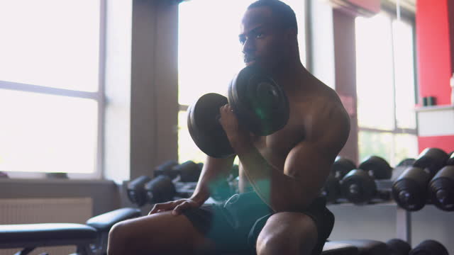 Muscular black man is working out in gym, lifting alternately big heavy dumbbell, doing exercises for biceps
