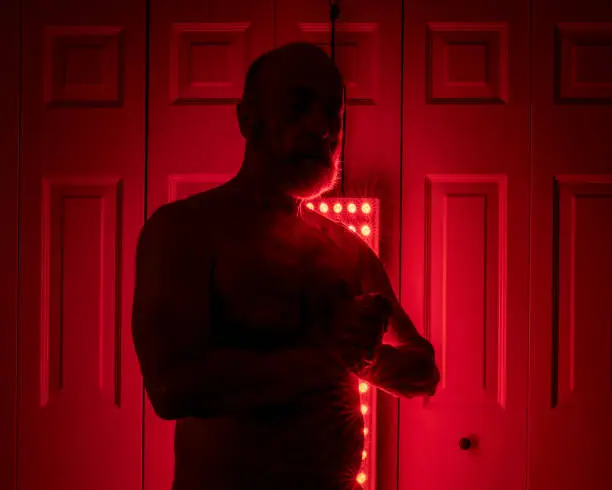 silhouette of a senior athletic, man torso standing in front of the red light therapy panel, home setup for treatment